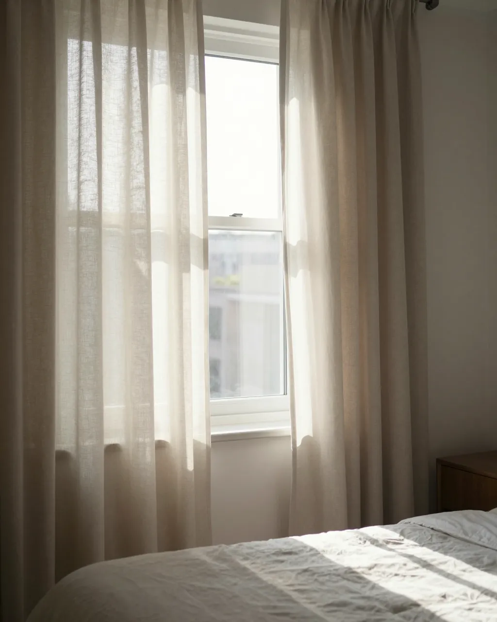 Soft morning sunlight streams through pulled-open linen curtains onto an unmade bedroom bed