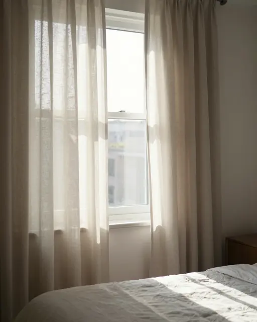 Soft morning sunlight streams through pulled-open linen curtains onto an unmade bedroom bed