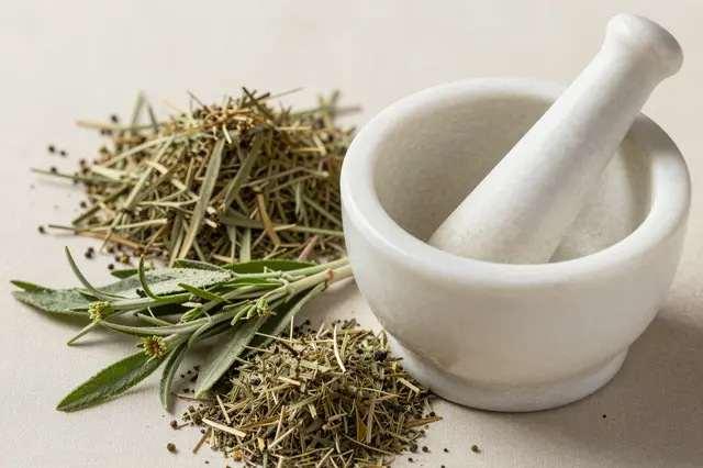 White marble mortar and pestle sit alongside fresh and dried medicinal herbs on a neutral light beige surface.