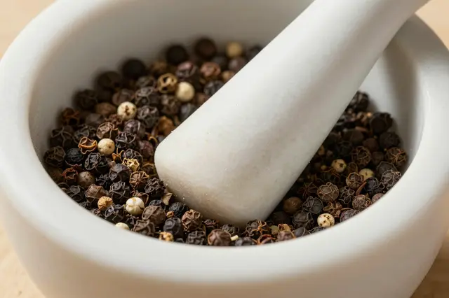 A close-up view of a white mortar and pestle filled with mixed whole peppercorns.