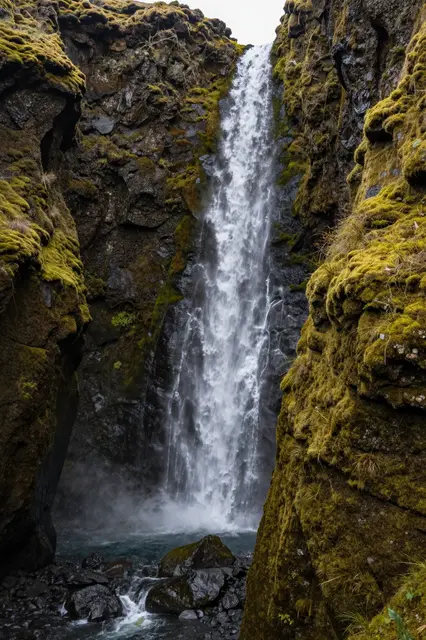 Tall narrow waterfall cascades between thick moss-covered rocky gorge walls into a dark pool below.