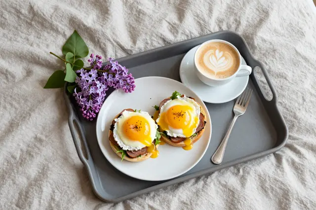 Overhead view of a bed tray with eggs Benedict, latte, and lilac sprigs on rumpled linen bedding