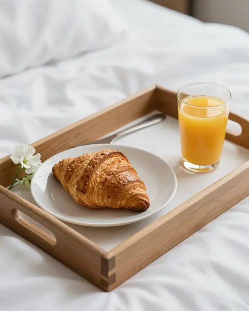 Wooden breakfast tray with croissant, orange juice and a sweet pea bloom rests on rumpled white bedding.