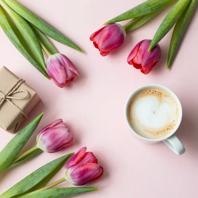 Top-down view of tulips, a small wrapped gift, and a cappuccino arranged on pale pink paper