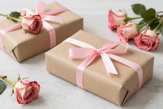 Wrapped kraft paper gift boxes with pink satin ribbon sit alongside pink roses on a light tabletop.