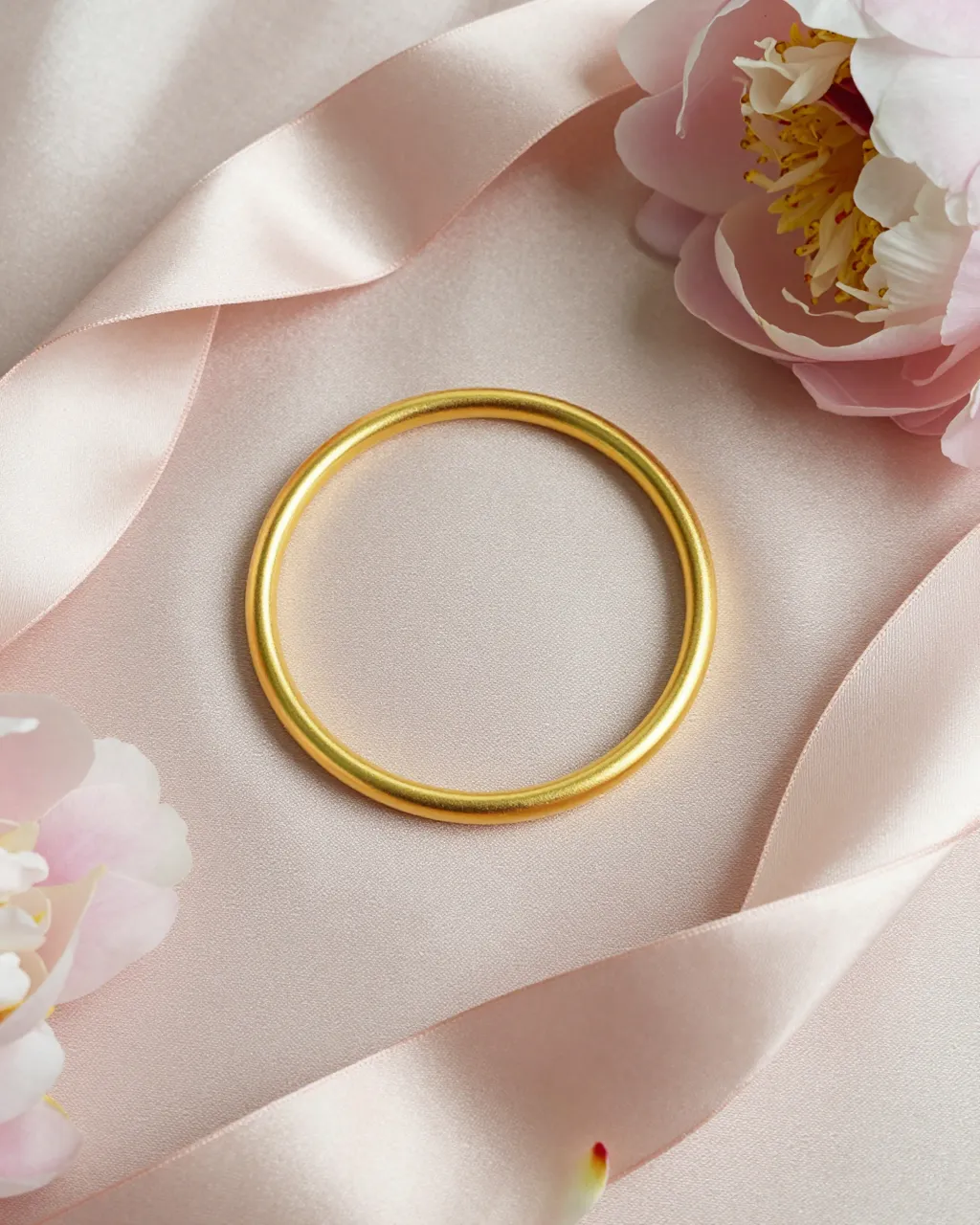 A polished gold bangle rests on blush satin surrounded by pink peonies and silk ribbon.