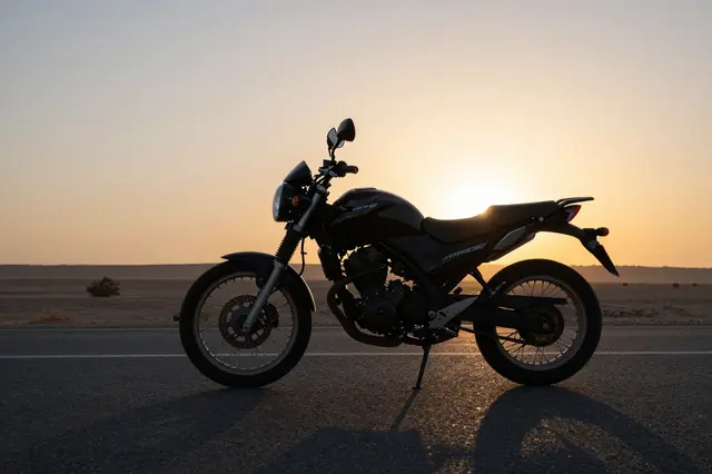 A parked motorcycle sits on an empty desert highway backlit by a glowing sunset sky.