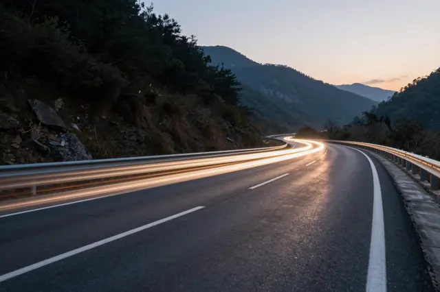 Long exposure light trails from passing vehicles curve along a winding mountain highway at twilight