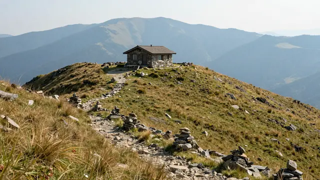 A stone mountain hut sits atop a grassy mountain saddle with stone cairns lining a hiking trail leading to distant hazy blue