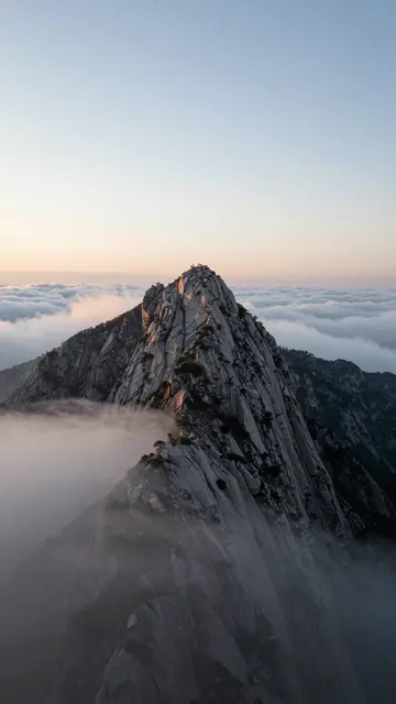 Jagged rocky mountain peak rises above a sea of soft clouds during pale warm sunrise