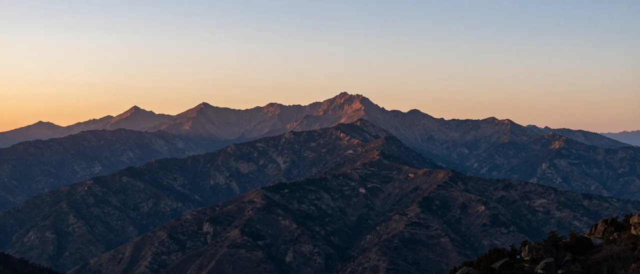 Layered mountain peaks stretch across the frame beneath a soft gradient sunset sky