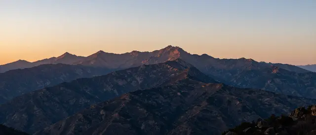 Layered mountain peaks stretch across the frame beneath a soft gradient sunset sky