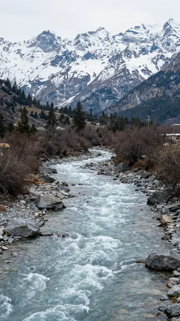 A fast flowing rocky turquoise mountain river leads toward tall snow covered alpine peaks under an overcast sky