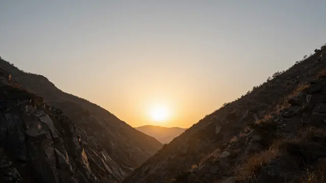 Bright sun sits between rocky mountain slopes casting warm golden light across the valley at sunset