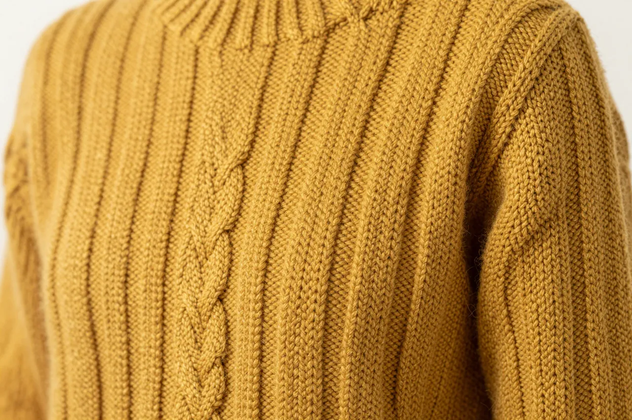 Close-up view of the thick braided cable knit texture of a mustard yellow wool sweater under soft side lighting
