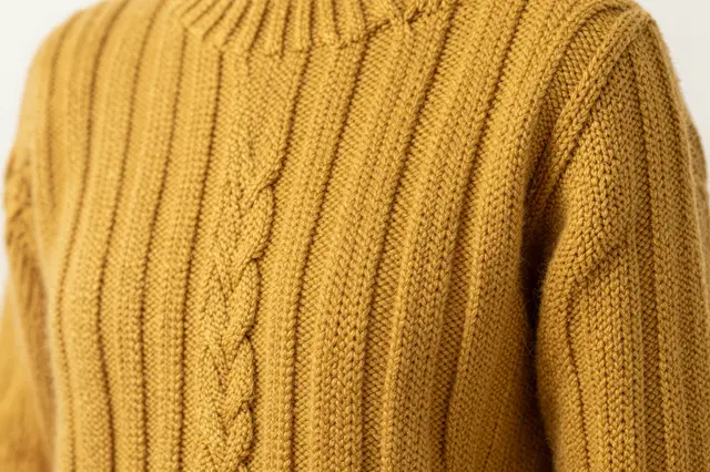 Close-up view of the thick braided cable knit texture of a mustard yellow wool sweater under soft side lighting