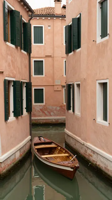 A wooden rowing skiff floats moored between ochre canal buildings with dark green shutters.