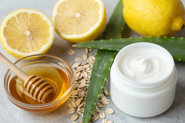 Flat lay arrangement of homemade skincare ingredients including lemons, honey, aloe, oats and cream jar on a gray surface