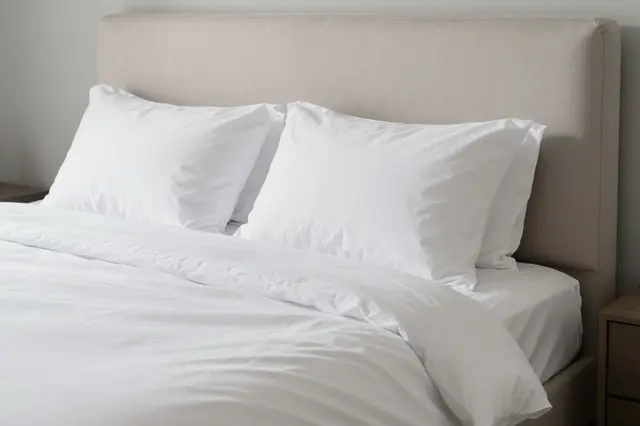 A neatly made bed with crisp white bedding sits against a light beige upholstered headboard in a minimalist bedroom.