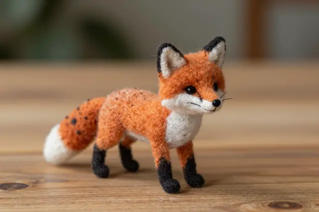 A small handmade needle-felted wool fox figurine stands on a light wooden table with a soft blurred background.