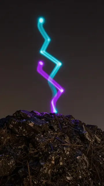 Cyan and magenta zigzag light trails arc above a dark rocky hilltop at night