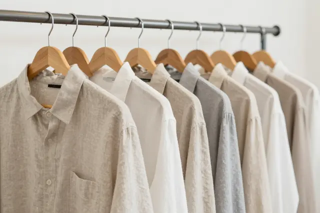 Row of plain neutral tone linen shirts hanging on wooden hangers in a bright minimalist interior