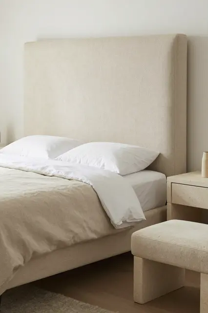 A minimalist primary bedroom with tall oatmeal linen headboard, stone nightstand, and upholstered bench at the bed foot