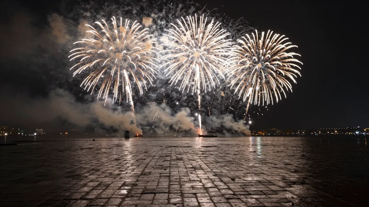 Golden white fireworks burst over wet cobblestone plaza reflecting light at midnight