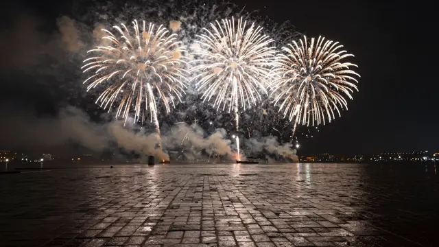 Golden white fireworks burst over wet cobblestone plaza reflecting light at midnight