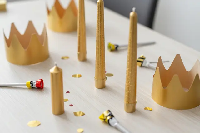 A light wooden party table arranged with gold paper crowns, golden candles, noisemakers and scattered confetti.