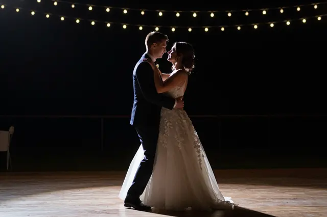 A bride and groom dance closely on a lit wooden dance floor at night under warm string lights