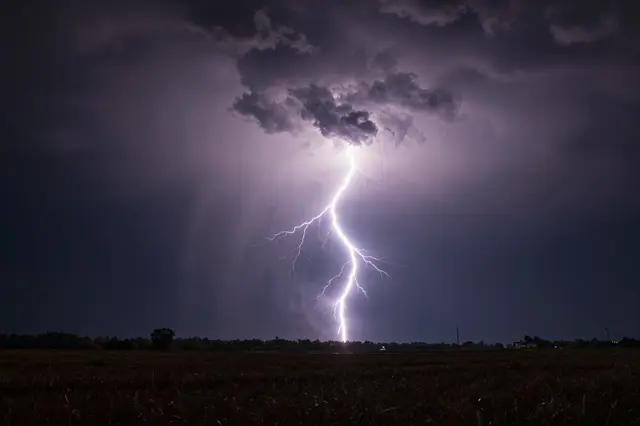 Bright lightning bolts erupt from a storm cloud over a dark rural field at night