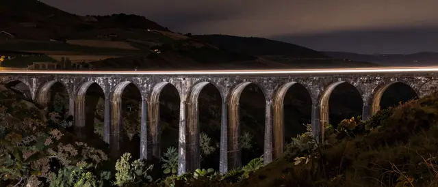 A continuous light streak crosses a stone arched viaduct in hilly countryside after dark