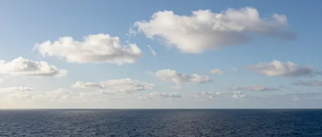 Calm dark blue ocean stretches to a sharp horizon under scattered fluffy white cumulus clouds in a bright pale blue noon sky.