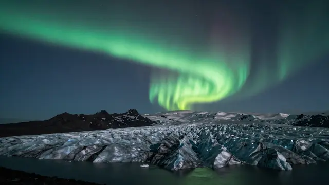 Swirling green aurora borealis arcs across a dark night sky above an Icelandic glacier lagoon