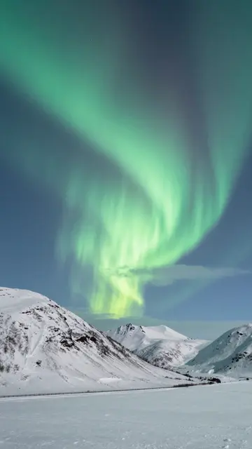 Swirling green northern lights arc across the sky above a snow-covered mountain range and frozen plain