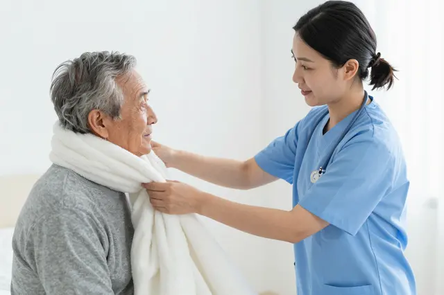 A female nurse in blue scrubs adjusts a soft white blanket around an elderly seated patient in a bright care room.