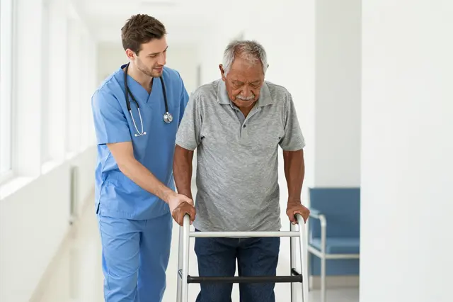 Young male nurse helps an elderly man use a walking frame in a bright hospital corridor.