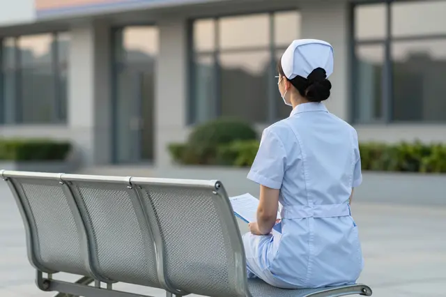 A nurse viewed from behind sits on an outdoor bench outside a hospital building holding a clipboard at dawn