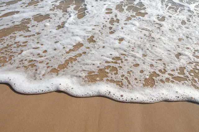 Top-down view of bubbly white ocean foam spreading across smooth tan beach sand