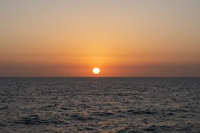 Bright round sun rests on the horizon above rippling dark ocean beneath a soft orange sunset sky