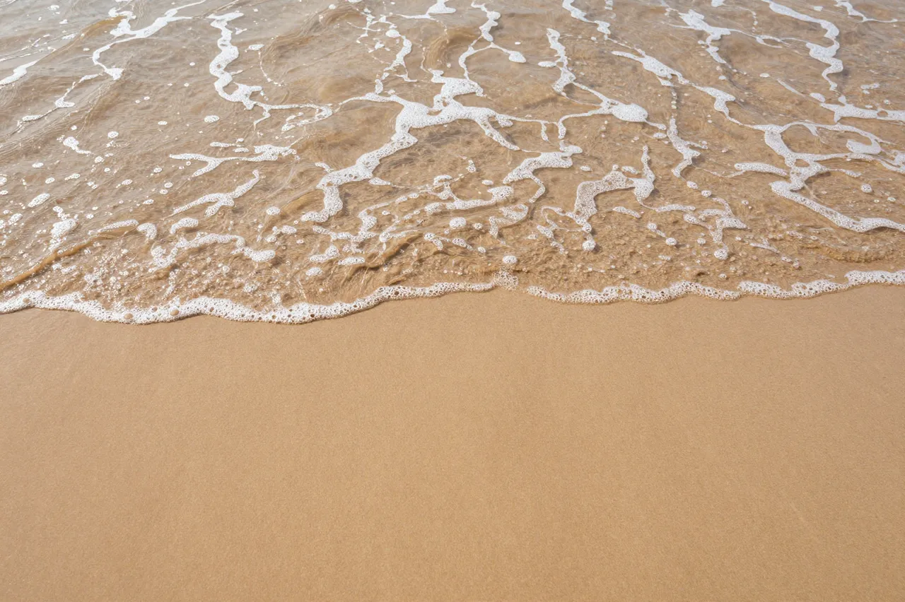 Top-down view of a foamy shallow ocean wave sliding over smooth tan beach sand