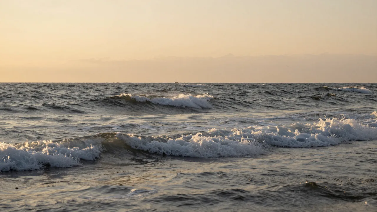 Rolling foamy ocean waves stretch across the frame beneath a hazy warm sunset sky