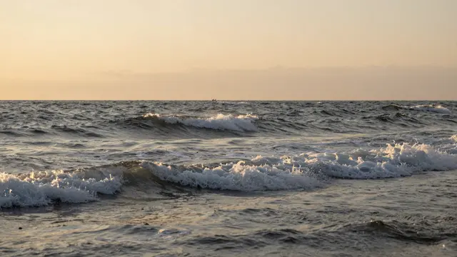 Rolling foamy ocean waves stretch across the frame beneath a hazy warm sunset sky