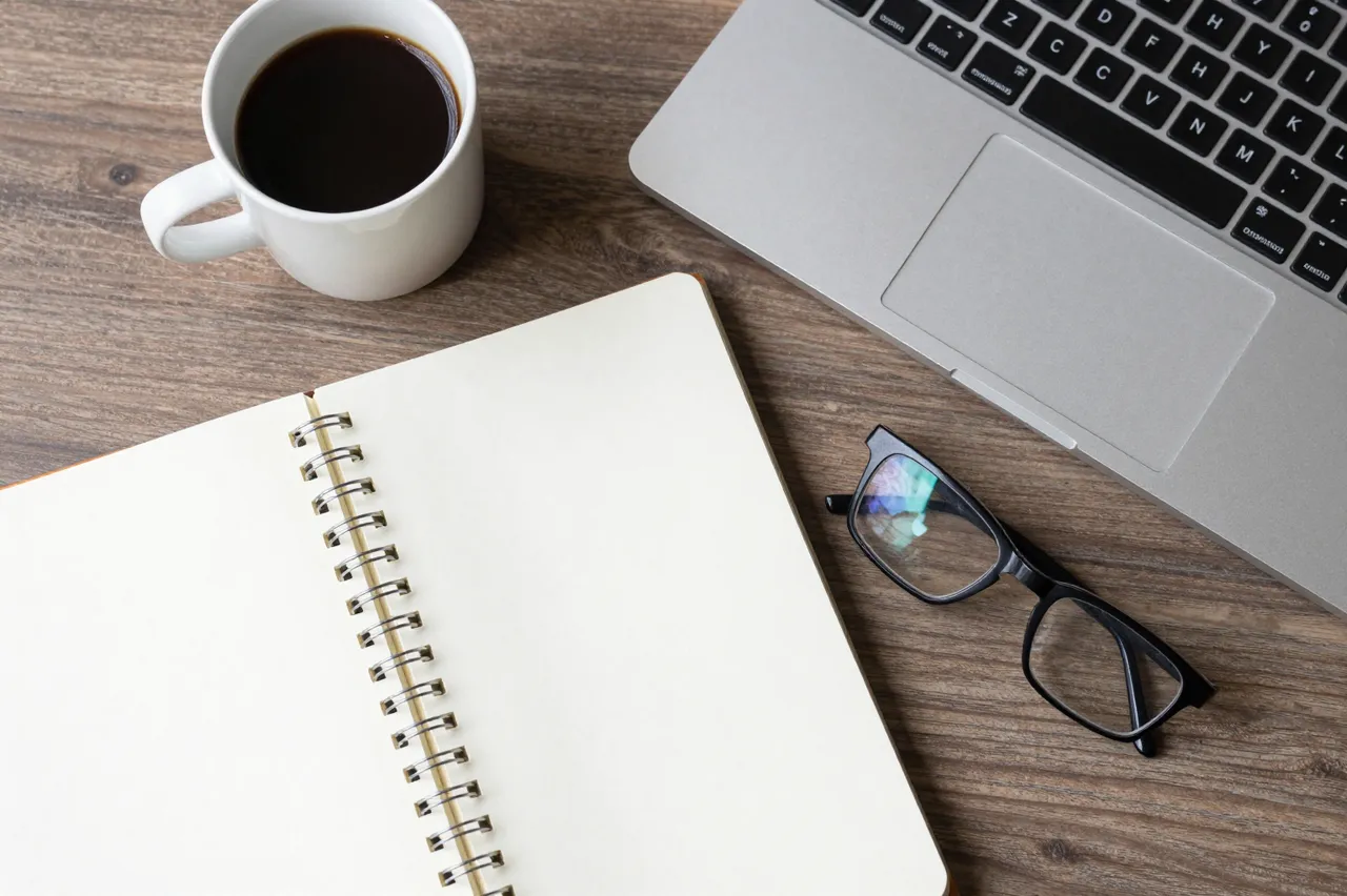 Top-down view of a wooden desk holding a notebook, coffee cup, eyeglasses, and silver laptop