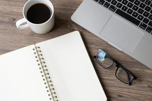 Top-down view of a wooden desk holding a notebook, coffee cup, eyeglasses, and silver laptop