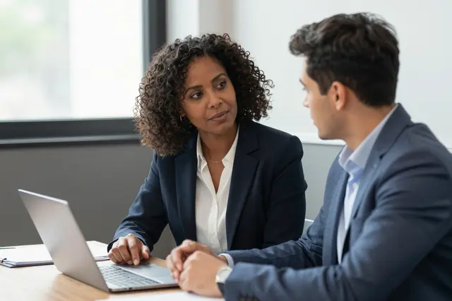 Two business colleagues converse across a laptop at an office conference table
