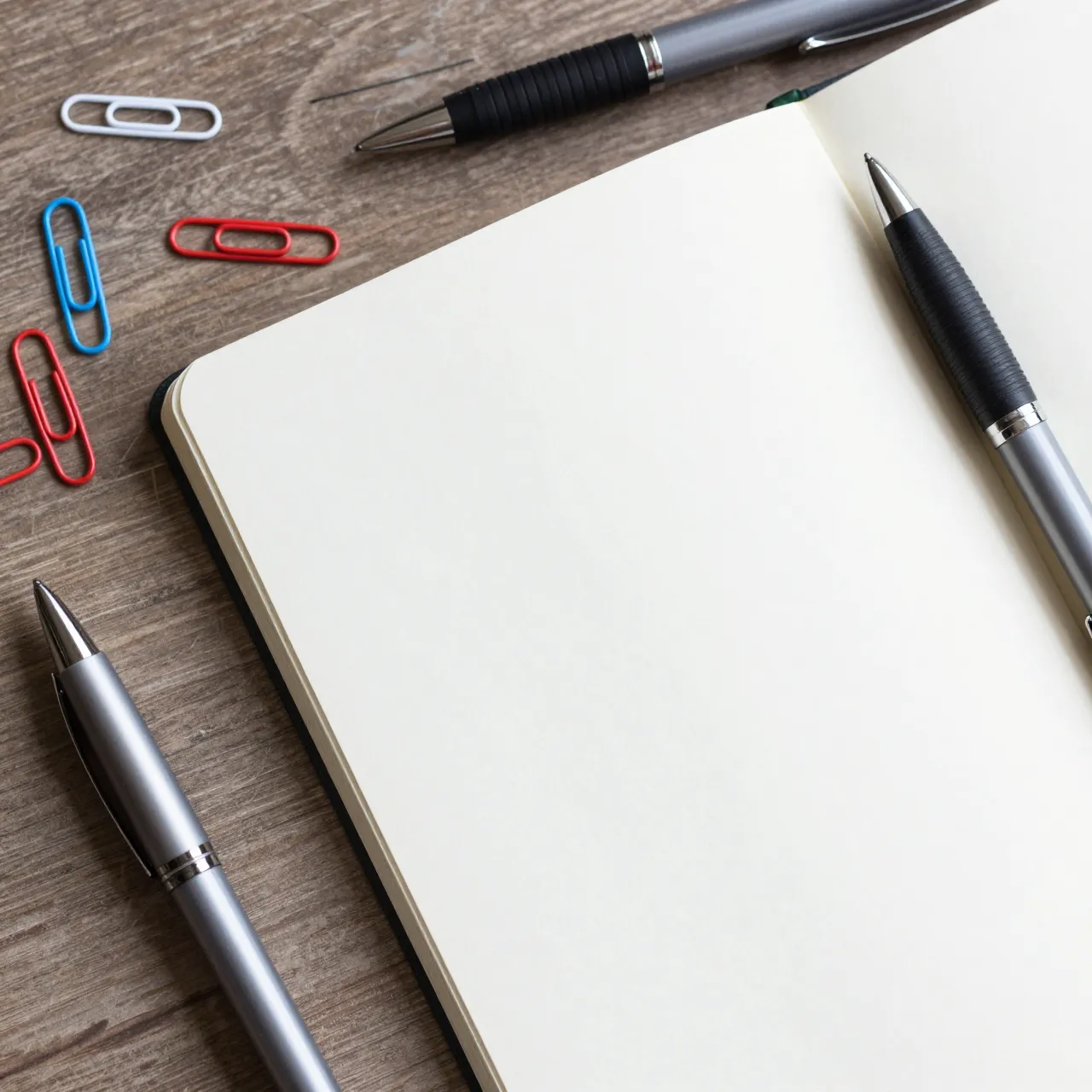 Top view of an open blank notebook, ballpoint pens and colorful paper clips on a wooden desk surface