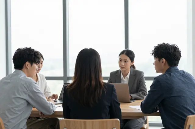 Four business colleagues sit around a wooden table in a bright office meeting near a large window