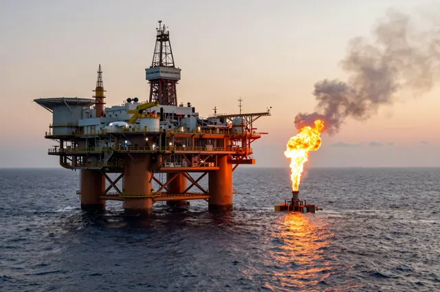 An offshore oil rig stands at sea beside a burning gas flare during warm sunset light